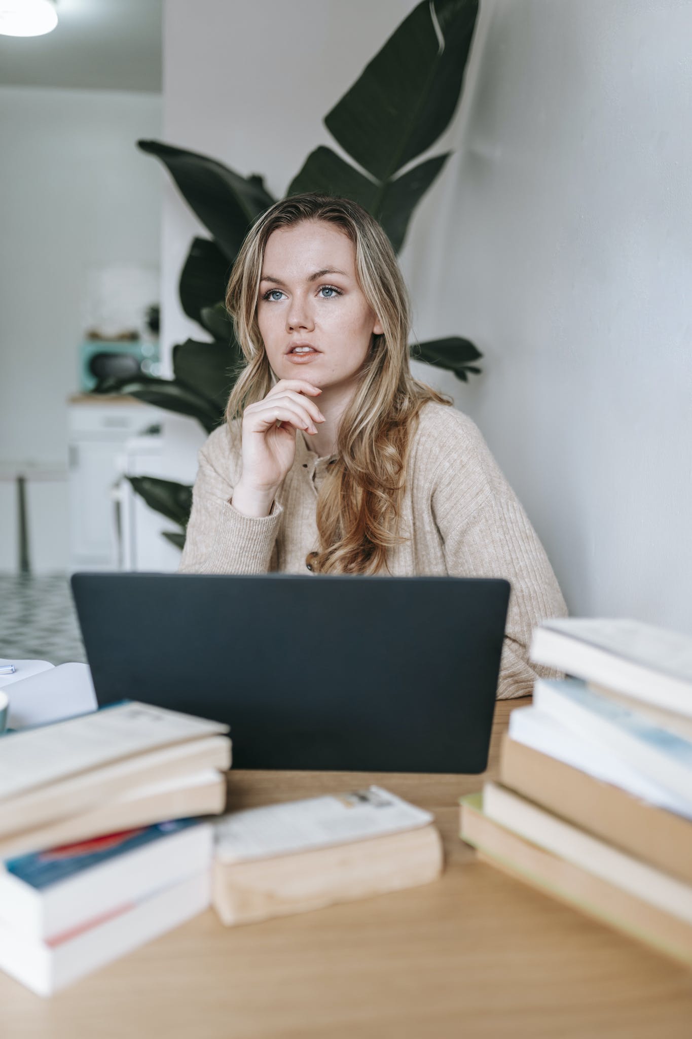 Pondering female using netbook near many books at table and green plant at home